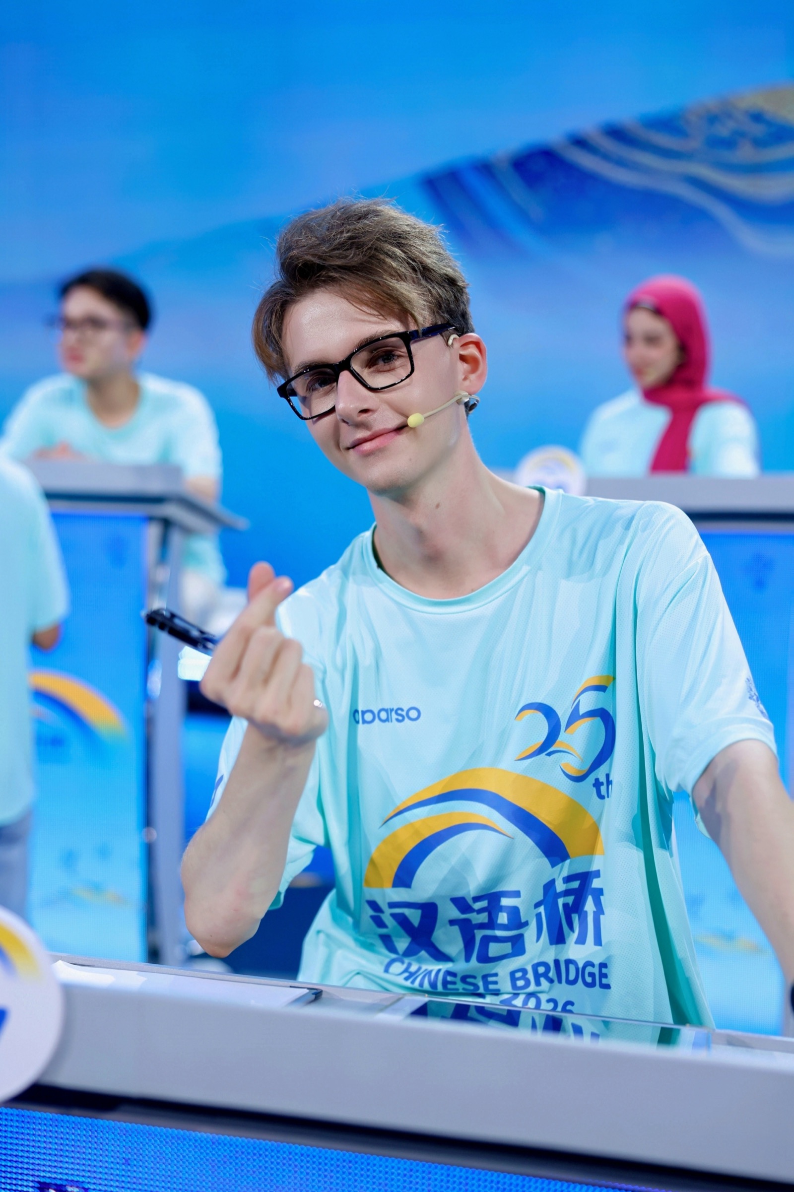 Levi im Chinese-Bridge-T-Shirt am Pult, lächelt in die Kamera und zeigt ein Fingerherz